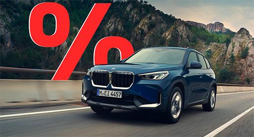  Das Bild zeigt einen dunkelblauen BMW X1, der auf einer Straße durch eine bergige Landschaft fährt. Auf der linken Seite befindet sich ein großes, rotes Prozentsymbol.