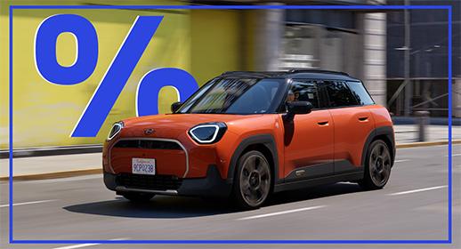  Das Bild zeigt einen orangefarbenen MINI Aceman Elektro, der auf einer Stadtstraße fährt. Das Auto hat ein schwarzes Dach und einen schwarzen Unterboden. Im oberen linken Teil des Bildes schwebt ein großes, blaues Prozentzeichen (%). Das Kennzeichen des Autos lautet "9CP238".