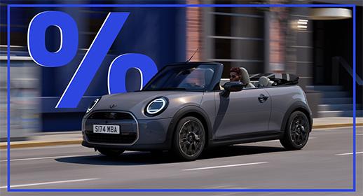 zeigt ein graues MINI Cooper Cabriolet, das auf einer Stadtstraße fährt. Das Verdeck ist geöffnet. Über dem Auto schwebt ein großes, blaues Prozentzeichen (%). Das Kennzeichen lautet "S I74 MBA".