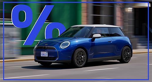 Dieses Bild zeigt einen blauen MINI Cooper Elektro 3-Türer, der auf einer Stadtstraße fährt. Das Auto hat ein weißes Dach und weiße Außenspiegel. Über dem Auto schwebt ein großes, blaues Prozentzeichen (%).