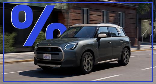  Das Bild zeigt einen grauen MINI Countryman mit Verbrennungsmotor, der auf einer Stadtstraße fährt. Das Auto hat ein beiges Dach. Über dem Auto schwebt ein großes, blaues Prozentzeichen (%). Das Kennzeichen lautet "9HK470".