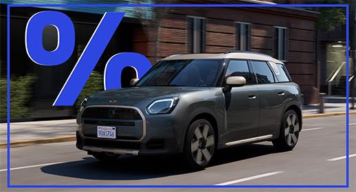  Das Bild zeigt einen grauen MINI Countryman Elektro, der auf einer Stadtstraße fährt. Das Auto hat ein beiges Dach. Über dem Auto schwebt ein großes, blaues Prozentzeichen (%). Das Kennzeichen lautet "9UA5746".