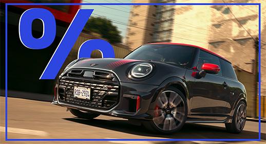 Das Bild zeigt einen grauen MINI Cooper im John Cooper Works (JCW) Design, der auf einer Straße fährt. Das Auto hat ein rotes Dach und rote Außenspiegel. Im oberen linken Bereich des Bildes ist ein großes, blaues Prozentzeichen (%) zu sehen. Das Kennzeichen lautet "R5B-2924".