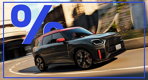 Das Bild zeigt einen grauen MINI Countryman im John Cooper Works (JCW) Design. Das Auto hat ein rotes Dach und rote Außenspiegel. Es fährt auf einer kurvigen Straße in einer städtischen Umgebung. Im oberen linken Bereich des Bildes schwebt ein großes, blaues Prozentzeichen (%). Das Kennzeichen lautet "D CK 58E".