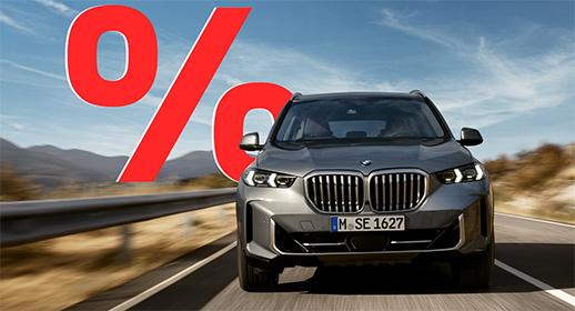 Das Bild zeigt ein graues SUV, den BMW X5, der auf einer Landstraße fährt. Das Auto ist von vorne zu sehen. Im Vordergrund, über dem Auto, ist ein großes, rotes Prozentzeichen (%) platziert. Der Hintergrund zeigt eine bergige Landschaft unter einem blauen Himmel mit einigen Wolken.