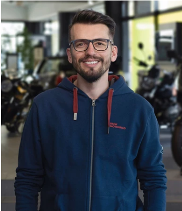 Das Bild zeigt einen Mann mittleren Alters, der direkt in die Kamera blickt und lächelt. Er hat einen kurzen Bart, trägt eine Brille mit dunklem Rand und einen dunkelblauen Kapuzenpullover. Auf der linken Brust des Pullovers ist in roter Schrift „BMW MOTORRAD“ zu lesen. Der Hintergrund ist unscharf und zeigt eine Motorrad-Ausstellungshalle mit mehreren Motorrädern.
