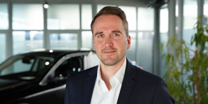 Ein professionelles Business-Porträt eines Mannes in dunklem Sakko und weißem Hemd, der freundlich in die Kamera blickt. Er steht in einem hellen, modern verglasten Raum, wobei ein Auto und eine Pflanze im Hintergrund dezent in Unschärfe verschwimmen. Die Aufnahme wirkt durch das weiche Licht besonders offen und kompetent.