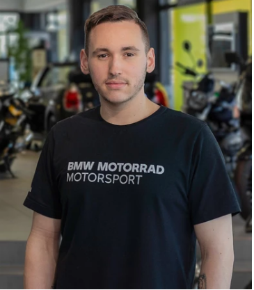 Das Bild zeigt einen jungen Mann mit kurzen, dunklen Haaren, der direkt in die Kamera blickt. Er trägt ein schwarzes T-Shirt, auf dem in weißen Buchstaben „BMW MOTORRAD MOTORSPORT“ und das BMW-Logo zu sehen sind. Der Hintergrund ist unscharf und zeigt eine Motorrad-Ausstellungshalle.