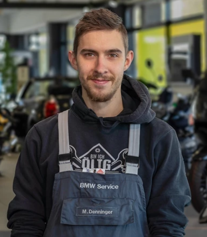 Ein junger Mann mit Bart und kurzen Haaren blickt in die Kamera. Er trägt einen grauen BMW Service-Overall über einem schwarzen Kapuzenpullover. Der Hintergrund ist eine unscharfe Autohaus-Kulisse.