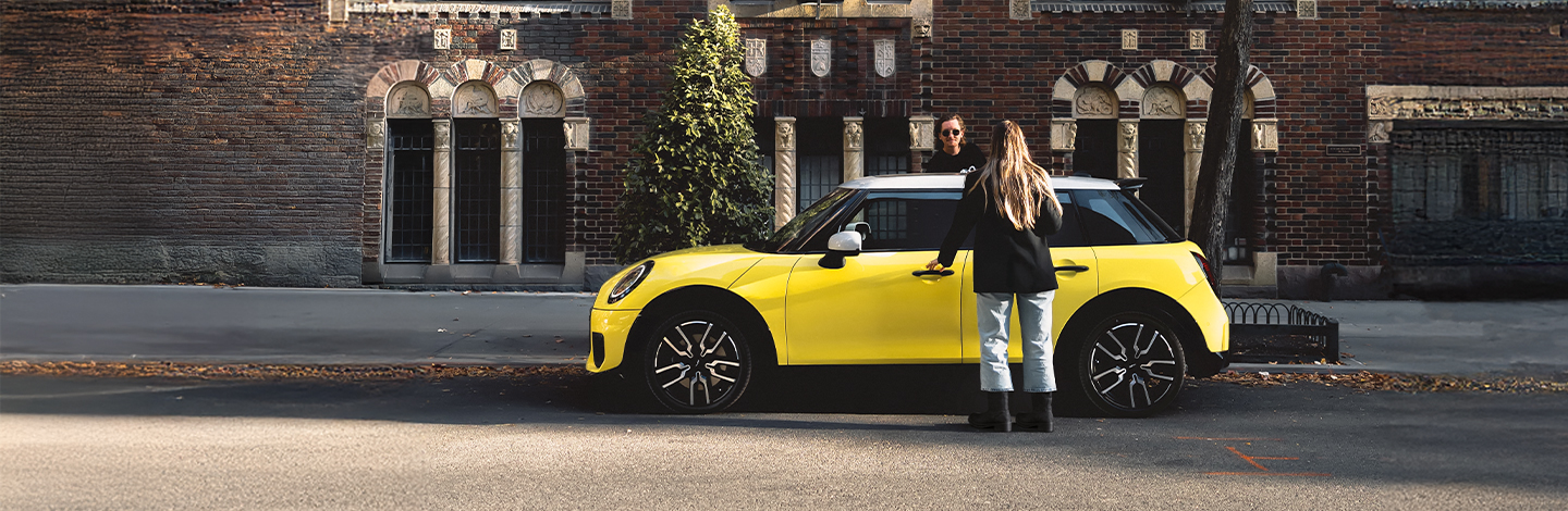 Das neue Bild zeigt einen leuchtend gelben Mini Cooper, der in einer städtischen Straße vor einer historischen Backsteinfassade parkt. Das Fahrzeug hat ein kontrastierendes weißes Dach und markante schwarze Felgen mit silbernen Details.  Eine Frau mit langem blondem Haar, gekleidet in einen schwarzen Blazer und helle Jeans, steht an der Fahrerseite und greift gerade nach dem Türgriff. Im Hintergrund sind verzierte, bogenförmige Fenster des Gebäudes und ein schmaler grüner Baum zu sehen. Die Szene ist in warmes Sonnenlicht getaucht, das lange Schatten auf den Asphalt wirft und dem Bild eine moderne Lifestyle-Atmosphäre verleiht.