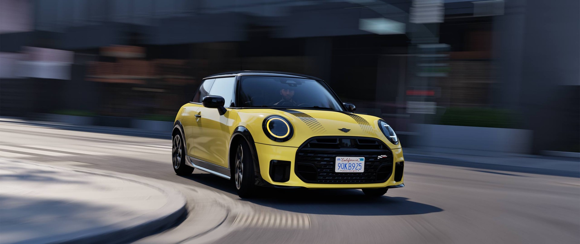 Ein gelber Mini Cooper mit schwarzem Dach und sportlichen Details fährt mit leichtem Bewegungsunschärfe-Effekt auf einer städtischen Straße. Das Auto ist dynamisch in einer Kurve abgebildet, was die Geschwindigkeit betont. Im Hintergrund sind unscharf Gebäude und ein grauer Himmel zu sehen. Das Fahrzeug trägt ein US-amerikanisches Nummernschild aus Kalifornien.