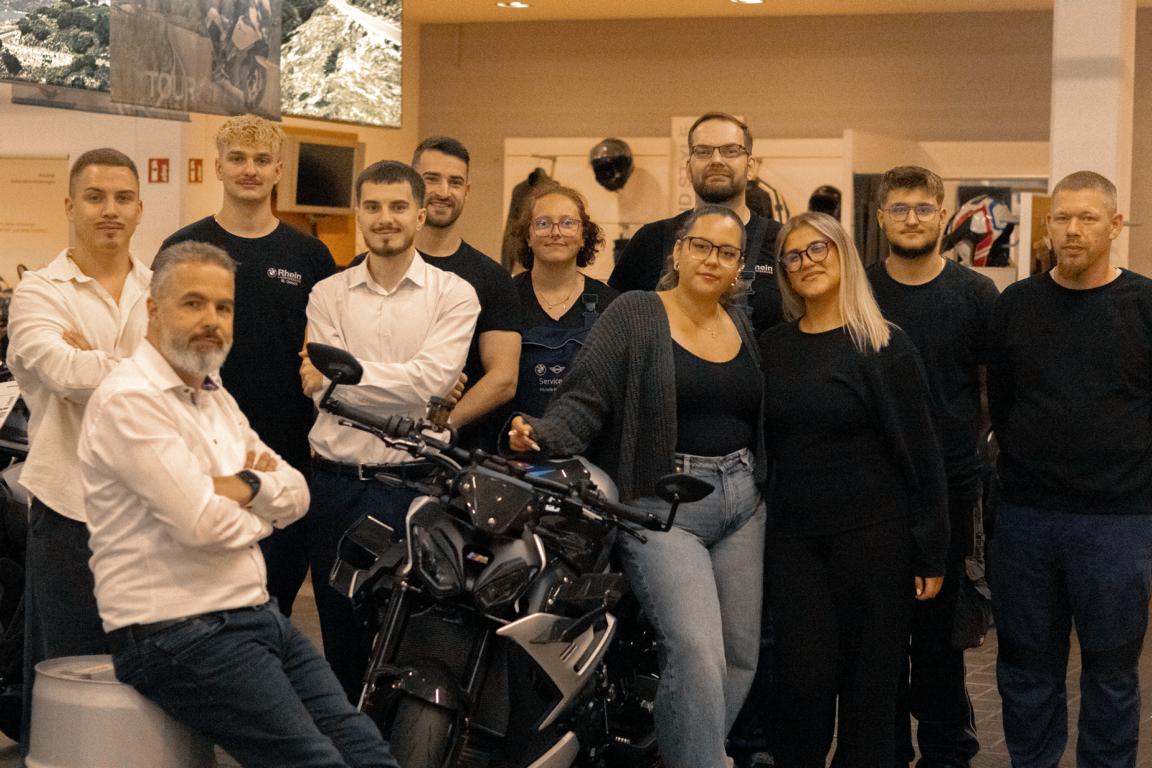 Das Bild zeigt ein Gruppenfoto eines zehnköpfigen Teams (sieben Männer und drei Frauen) in einem Motorrad- oder Autohaus. Im Mittelpunkt der Gruppe steht ein sportliches, dunkles BMW-Motorrad
