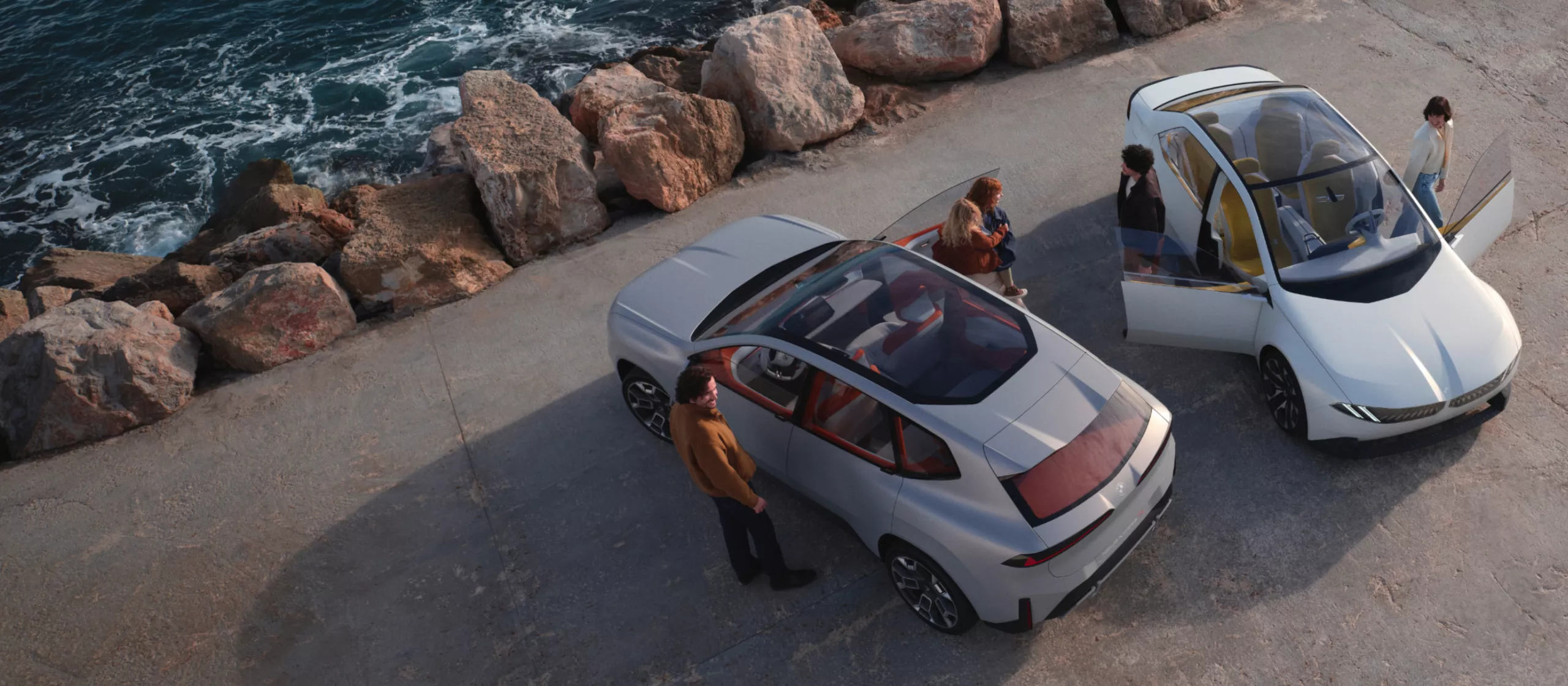 Das Bild zeigt eine Szene am Meer, in der zwei futuristische Elektroautos stehen. Im Hintergrund ist das Meer zu sehen, dessen Wellen an einen felsigen Küstenstreifen schlagen. Im Vordergrund stehen zwei Autos nebeneinander auf einem asphaltierten Weg. Beide sind weiße SUVs mit großen, gläsernen Dachflächen. Die Türen der Autos sind geöffnet, und in und um die Autos herum stehen mehrere Menschen. Sie scheinen miteinander zu interagieren und die Fahrzeuge zu betrachten.
