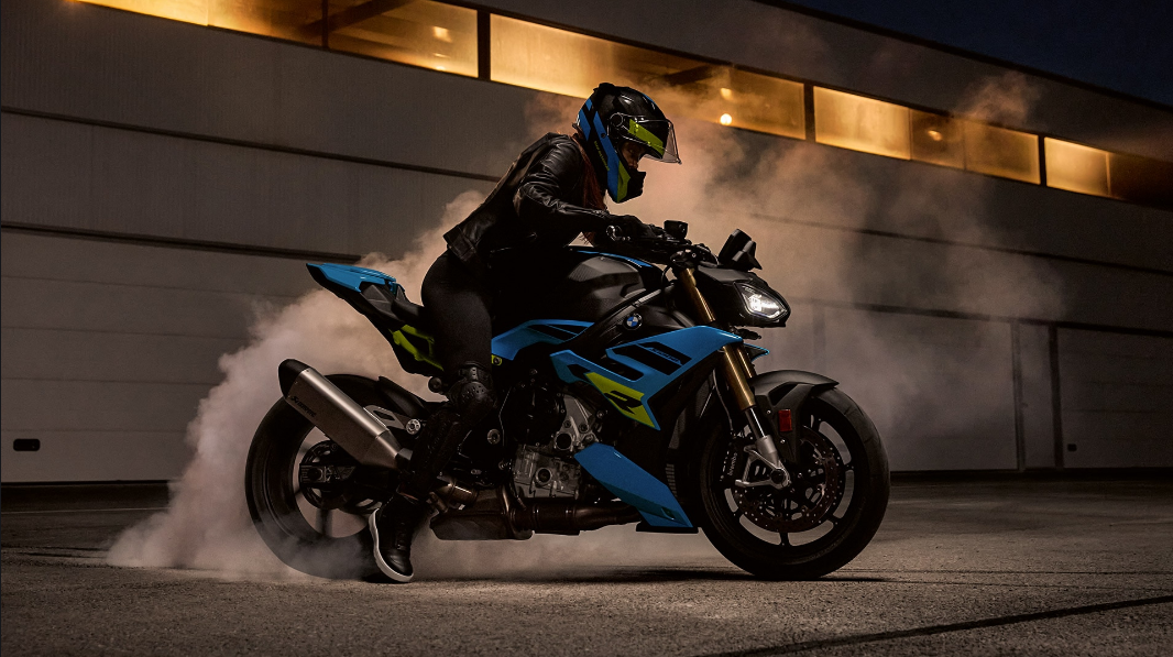 Eine Motorradfahrerin in dunkler Lederjacke, Leggings und einem blau-gelben Helm fährt ein türkisfarbenes BMW Naked Bike auf einer dunklen Fläche, während Rauch oder Nebel um das Hinterrad aufsteigt. Das Motorrad ist in einem auffälligen Türkisblau und Schwarz gehalten und hat neongelbe Akzente. Die Fahrerin ist seitlich aufgenommen, wie sie sich dynamisch nach vorne beugt. Im Hintergrund leuchten rechteckige Fenster eines Gebäudes in warmem Gelb. Die Szene ist dramatisch und vermittelt eine sportliche, nächtliche Fahrt.