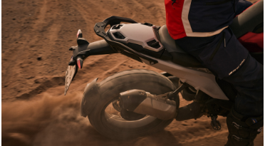 Nahaufnahme des Hinterrads eines Motorrads, das auf einem staubigen, trockenen Boden fährt und dabei Sand aufwirbelt. Die Aufnahme ist von der Seite und zeigt das Hinterrad mit einem schwarzen Schutzblech, das in einer dynamischen Staubwolke verschwindet. Die Aufhängung ist teilweise sichtbar, ebenso wie Teile des Auspuffs und die rote Verkleidung des Fahrers. Die Szene fängt die Dynamik des Fahrens im Gelände ein.