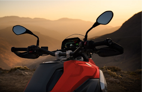 Ein rotes BMW Adventure-Motorrad steht bei Sonnenuntergang auf einem Bergkamm und blickt über eine weite, neblige Hügellandschaft. Das Bild ist aus der Perspektive des Fahrers aufgenommen und zeigt das Cockpit des Motorrads mit einem eingeschalteten, digitalen Display, das den Leerlauf "N" anzeigt. Der Lenker ist mit schwarzen Handschützern und zwei Rückspiegeln ausgestattet. Das warme, goldene Licht der untergehenden Sonne scheint über die in sanfte Schatten getauchten Berge im Hintergrund. Das Bild vermittelt eine ruhige und majestätische Atmosphäre.