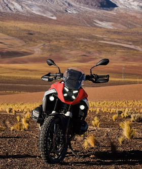 Ein rotes und weißes BMW Adventure-Motorrad steht auf einer kargen, sandigen Ebene, die von großen, braunen Bergen umgeben ist. Das Motorrad ist von vorne aufgenommen und seine markanten LED-Scheinwerfer leuchten hell. Im Vordergrund sind vereinzelt trockene Grasbüschel zu sehen. Im Hintergrund ragen gewaltige Berge auf, deren obere Gipfel mit weißem Schnee bedeckt sind. Die gesamte Szene vermittelt ein Gefühl von Abenteuer und rauer, unberührter Natur.
