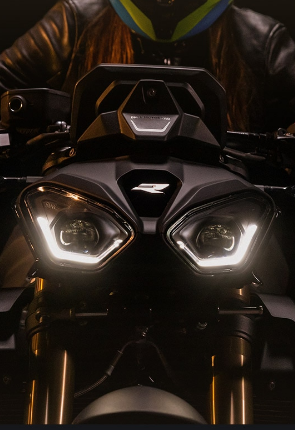 Das Bild zeigt die Front eines BMW-Motorrads im Dunkeln. Die markanten LED-Doppelscheinwerfer leuchten hell und geben dem Motorrad einen kraftvollen, fokussierten Ausdruck. Dahinter ist ein Fahrer oder eine Fahrerin mit Helm und Lederjacke erkennbar.