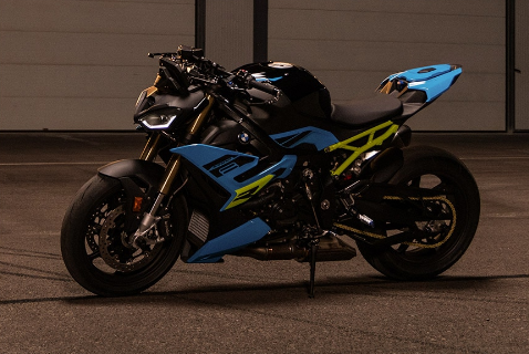 Auf dem Bild ist ein sportliches BMW-Motorrad zu sehen, das in kräftigem Blau mit gelben Akzenten lackiert ist. Es steht aufrecht mit Seitenständer in einer Halle oder auf einem asphaltierten Platz. Das Design wirkt dynamisch, aggressiv und modern.