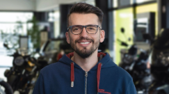 Leo Deda - Verkaufsleiter Motorrad Heermann Rhein Heilbronn.  Auf dem Bild ist eine Porträtaufnahme eines Mannes zu sehen, der in einem Motorradgeschäft steht.  Der Mann schaut direkt in die Kamera, trägt eine Brille und hat einen leichten Bart. Er lächelt freundlich und ist in einen blauen Kapuzenpullover gekleidet. Der Hintergrund ist unscharf, man kann aber Motorräder und Teile des Geschäfts erkennen.