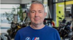 Dejan Sarcevic Verkaufsberater Motorrad Heermann Rhein Heilbronn.  Auf dem Bild ist eine Porträtaufnahme eines Mannes zu sehen, der in einem Motorradgeschäft steht.  Der Mann schaut direkt in die Kamera, trägt ein blaues T-Shirt mit einem BMW M-Logo und hat graues, kurzes Haar. Er lächelt freundlich. Der Hintergrund ist unscharf, man kann aber Motorräder und Teile des Geschäfts erkennen.