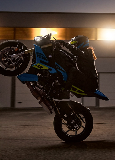 Auf dem Bild ist ein Motorradfahrer oder eine Motorradfahrerin zu sehen, der auf einem BMW-Motorrad einen spektakulären Wheelie macht – das Vorderrad ist hoch in der Luft. Die Szene wirkt dynamisch, sportlich und findet in der Dämmerung vor einer Halle mit hellen Lichtern statt.