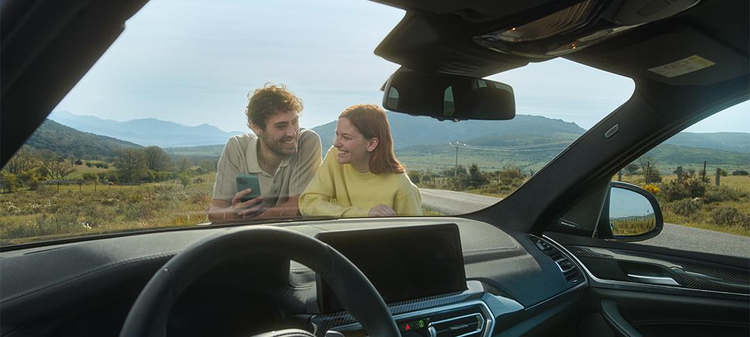 BMW Service Inclusive : Aus der Perspektive des Fahrzeuginnenraums blickt man durch die Windschutzscheibe auf ein lächelndes Paar, das sich von außen an die Scheibe lehnt. Der Mann hält ein Smartphone. Sie stehen am Straßenrand in einer ländlichen, hügeligen Landschaft.