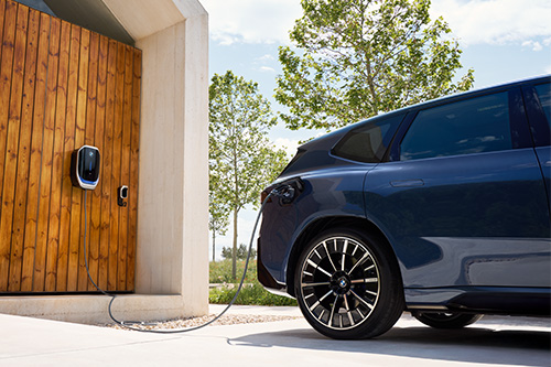 Die Aufnahme konzentriert sich auf die Heckpartie eines dunkelblauen SUV während des Ladevorgangs. Das Auto parkt auf hellem Beton. Das Ladekabel ist in der rechten hinteren Flanke des Wagens eingesteckt. Das Kabel ist mit einer schwarzen Wallbox verbunden, die an einer großen Fläche aus vertikalen Holzlatten (vermutlich einer Garagentür) montiert ist. Im Hintergrund erzeugen üppiges Grün und hohe Bäume an einem sonnigen Tag eine frische, ruhige Umgebung.