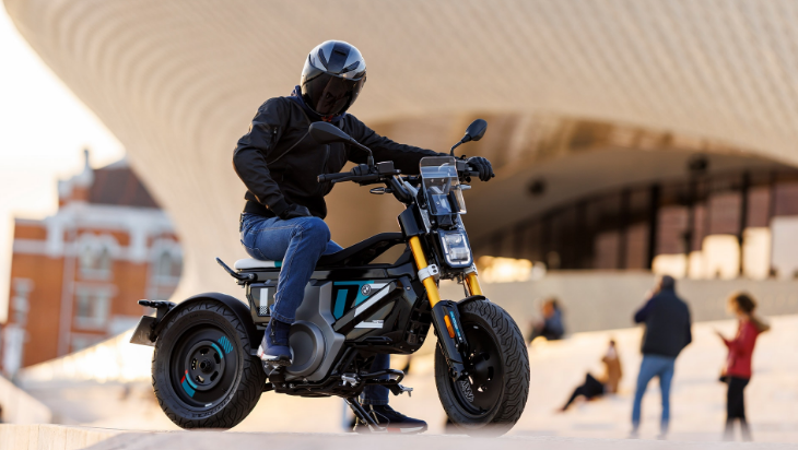 Ein Fahrer in schwarzer Jacke, blauen Jeans und einem geschlossenen, schwarzen Motorradhelm sitzt auf einem kompakten, futuristisch aussehenden Elektroroller, der vor einer modernen, geschwungenen Architektur steht. Der Roller hat dicke Reifen, eine gelbe Federung und ein kastenartiges Design des Motors in Schwarz, Grau und Türkis. Die Szene ist in warmes Abendlicht getaucht. Im unscharfen Hintergrund sind weitere Personen zu erkennen, die sich auf einer Terrasse oder einem Platz aufhalten.