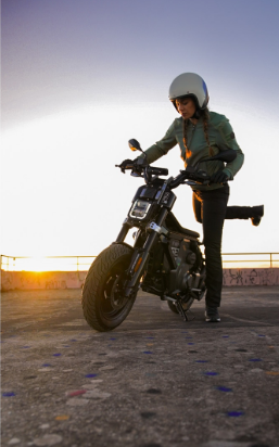 Eine junge Frau mit langen Zöpfen, bekleidet mit einer olivgrünen Jacke und schwarzen Hosen, steht neben einem mattschwarzen Motorrad mit robusten Reifen. Sie trägt einen weißen Helm und legt gerade ihre rechte Hand an den Lenker. Die Szene ist von einem tiefstehenden Sonnenuntergang beleuchtet, dessen oranges Licht am Horizont, direkt hinter dem Motorrad, stark leuchtet. Die Aufnahme entstand auf einem leicht verschmutzten, grauen Asphaltboden, der mit verblichenen Farbmarkierungen übersät ist.