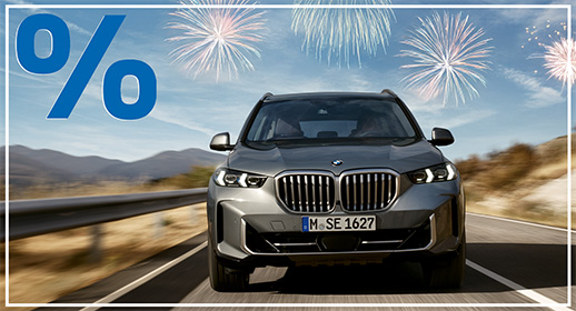 Dieses Bild präsentiert einen massiven, grauen BMW X5 SUV in einer direkten Frontansicht, während er über eine asphaltierte Straße fährt. Die Umgebung wirkt weitläufig mit Bergen im Hintergrund und einer Leitplanke am Straßenrand, was ein Gefühl von Freiheit und Abenteuer vermittelt. Der blaue Himmel wird von mehreren bunten Feuerwerken geschmückt, während das große blaue Prozentzeichen oben links und der weiße Außenrahmen das Design der Aktionsserie vervollständigen.